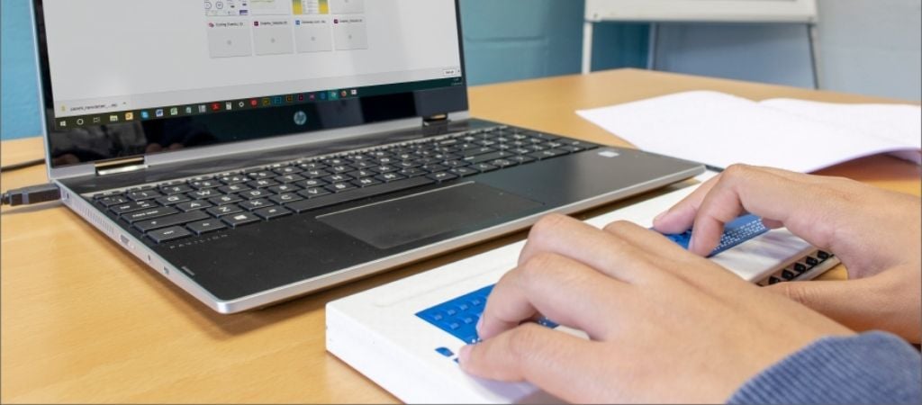 Open laptop in front of a braille display being used.  The image shows someones hands as they read with the braille display